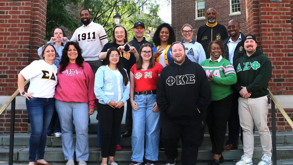 A large group of staff stand outside. They are all wearing Greek letters.